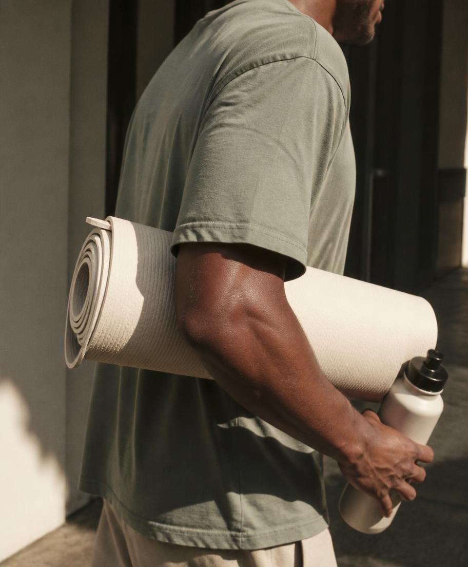 Close crop of a man leaving a workout with a rolled cream mat under his arm and a water bottle in hand, warm sunlight across his skin