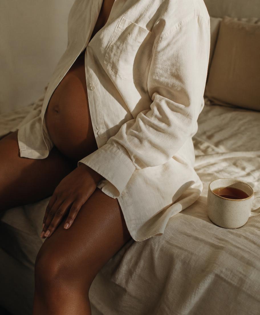 Intimate crop of a pregnant woman in an oversized cream linen shirt sitting on rumpled bed linen beside a mug of tea in morning light